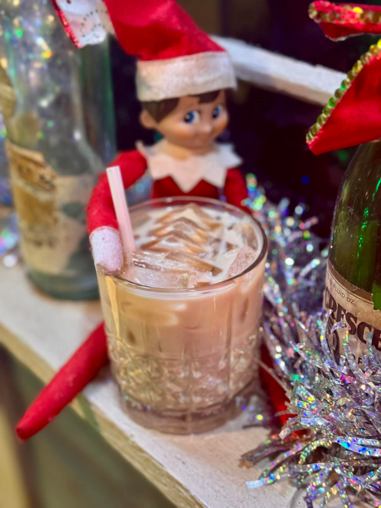 An Elf on the Shelf perched beside a festive holiday cocktail.