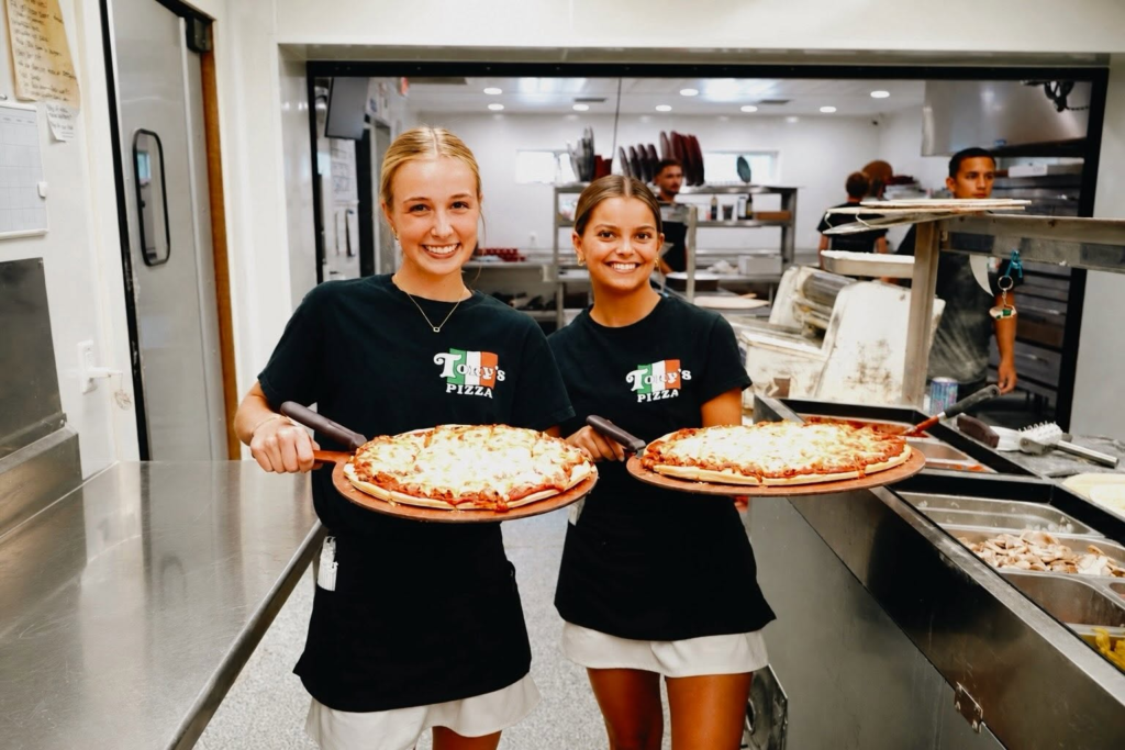 Two smiling staff members holding fresh pizzas at a local pizzeria.