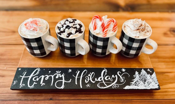 A holiday coffee flight in black and white plaid cups on a Happy Holidays board.